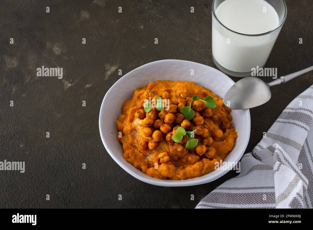 Dicke Kürbissuppe mit gebackenen Kichererbsen und Rettichsprossen auf einem Teller mit einem Glas Milch auf dem Tisch, leckeres gesundes Abendessen Stockfoto