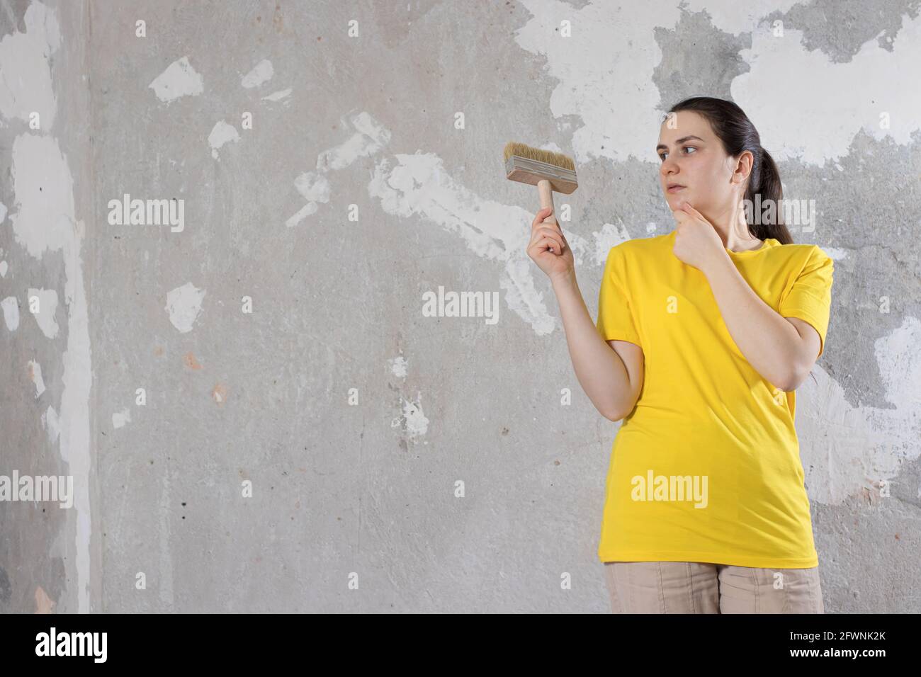 Eine Reparaturfrau in einem gelben, hellen T-Shirt hält einen Pinsel und blickt auf eine leere Wand mit Platz für Text. Schlüsselfertige Reparaturen. Stockfoto