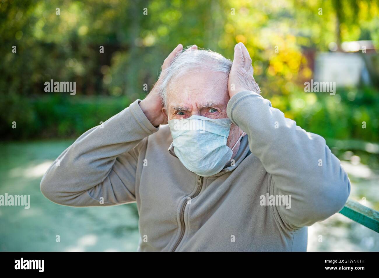 Kranker, trauriger grauhaariger alter Mann sitzt in einer medizinischen Maske auf der Straße. Der Mann hat starke Kopfschmerzen. Das Konzept der Kopfschmerzen. Frühjahrsallergie, Methoden der Stockfoto