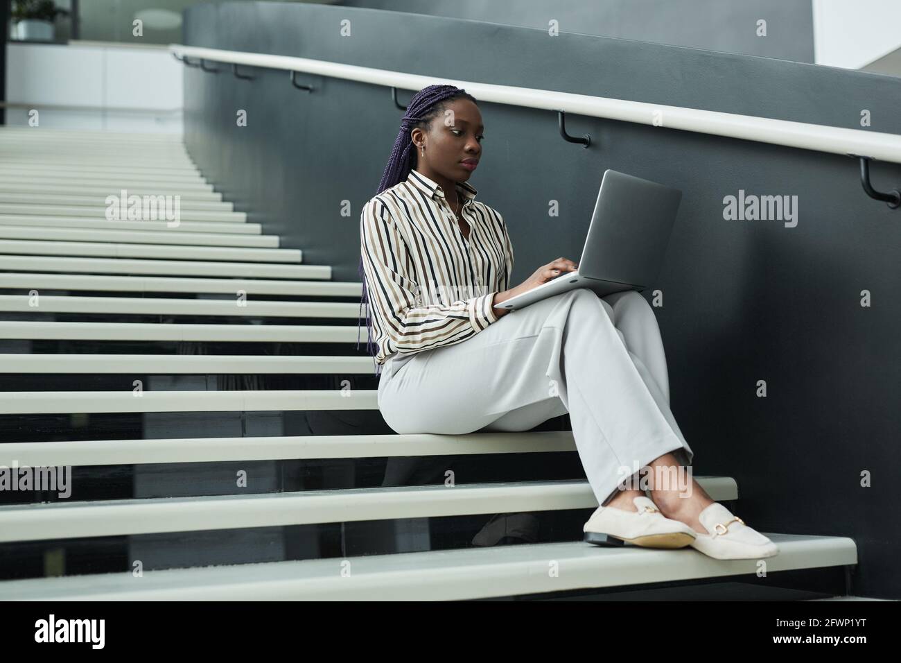 Grafisches Porträt einer jungen afroamerikanischen Geschäftsfrau in Schwarz-Weiß mit Laptop, während sie im Büro auf einer Treppe sitzt, Kopie sp Stockfoto