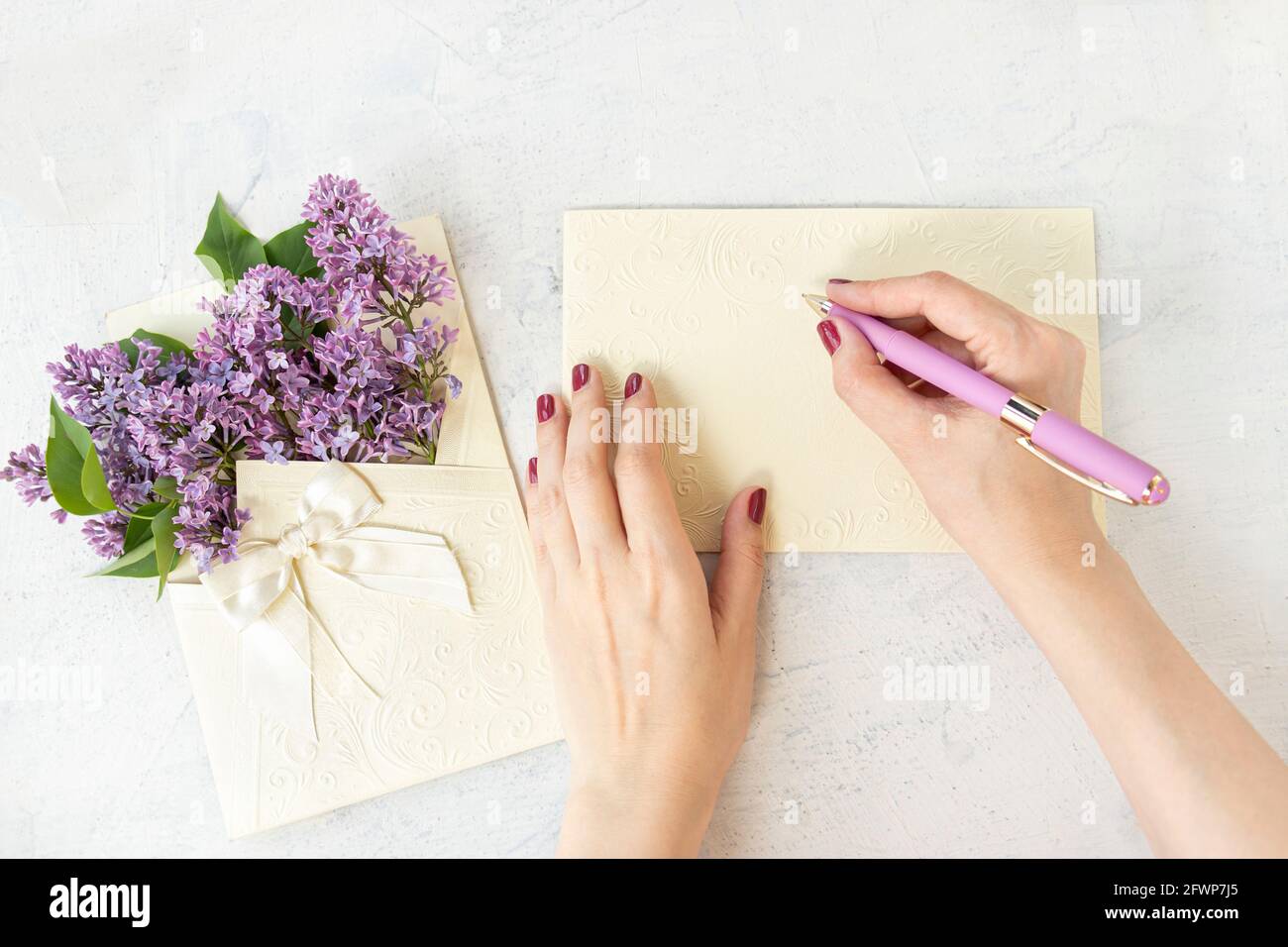 Die Hände der Frau schreiben auf einer leeren Karte oder leeren, in der Nähe von violetten Blumen in einem schönen Umschlag mit einer Schleife. Wunderschöne Fliederblüten. Flach legen mit Feder blo Stockfoto