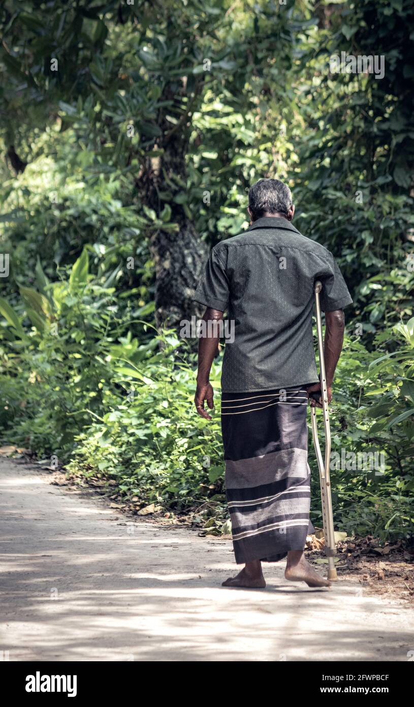 Ältere Erwachsene gehen mit Hilfe eines Stockes in einer einsamen ländlichen Dorfstraße. Stockfoto