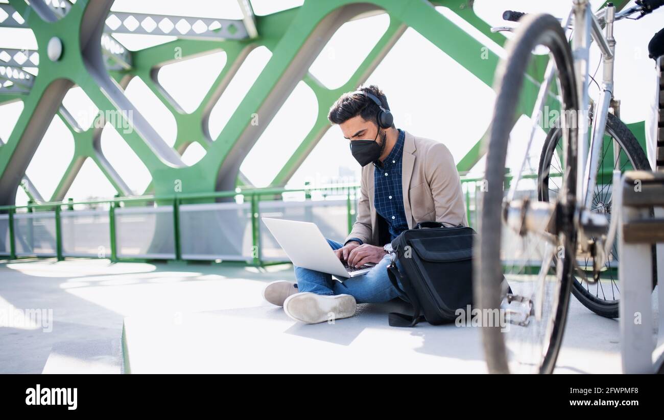 Der junge Geschäftsmann Pendler mit Laptop arbeitet auf dem Weg zur Arbeit im Freien auf der Brücke in der Stadt, Coronavirus-Konzept. Stockfoto
