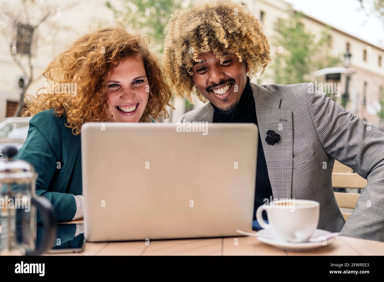 Multikulturelle Geschäftsleute, die draußen in einer Cafeteria sitzen und Kaffee trinken und mit einem Laptop arbeiten. Sie lächeln und schauen auf den Computerbildschirm Stockfoto