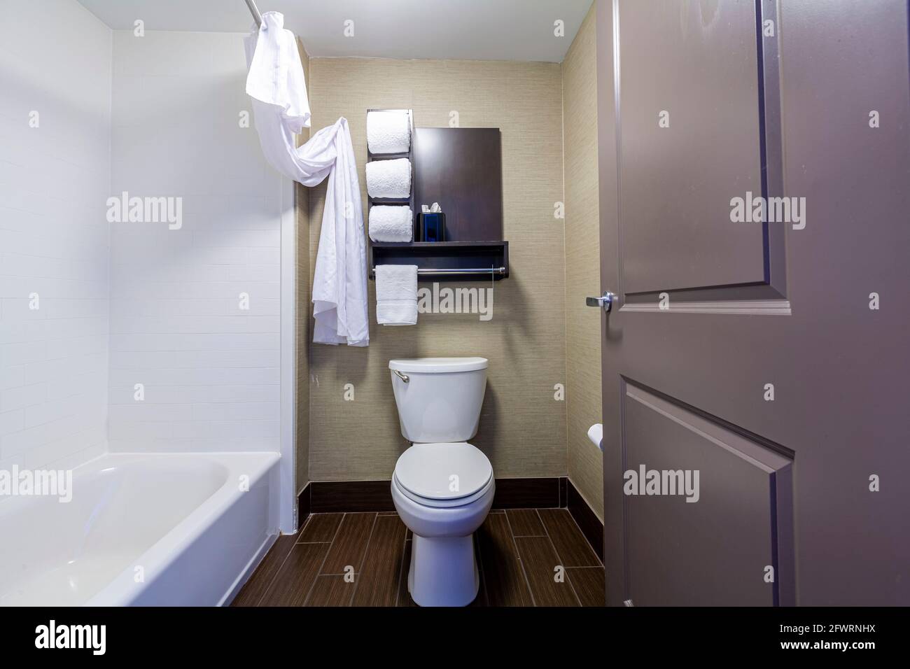 Ein kleines Badezimmer und Toilette in einem Hotel mit weißen Handtüchern gefaltet und auf Regalen platziert. Eine Badewanne mit Duschvorhang befindet sich auf der linken Seite. Ein minimalistischer De Stockfoto