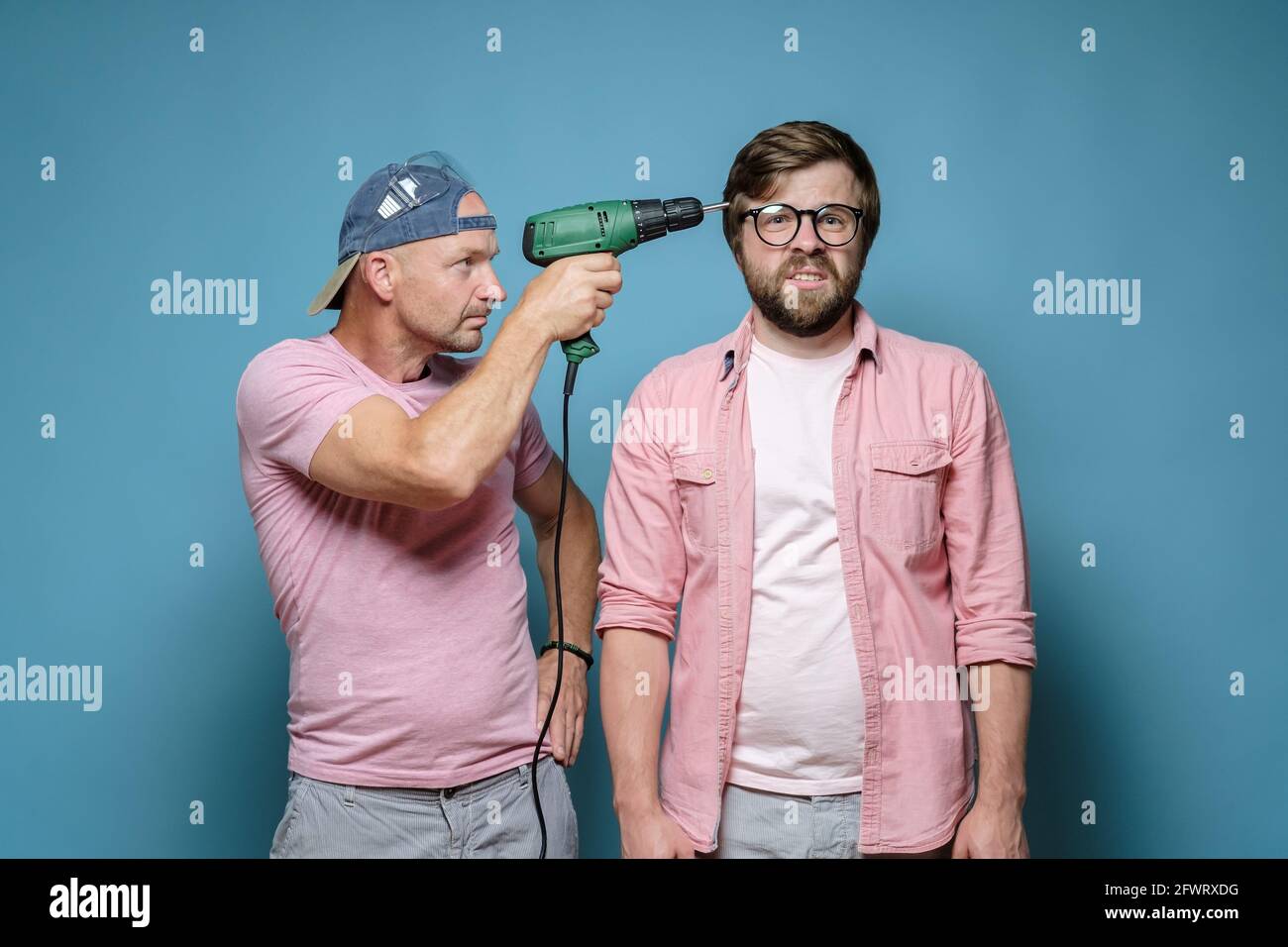 Nachbar macht Reparaturen. Ein ernsthafter Mann steckte einen elektrischen Schraubendreher an den unglücklichen männlichen Kopf. Lärm aus dem Haushalt. Stresskonzept. Stockfoto