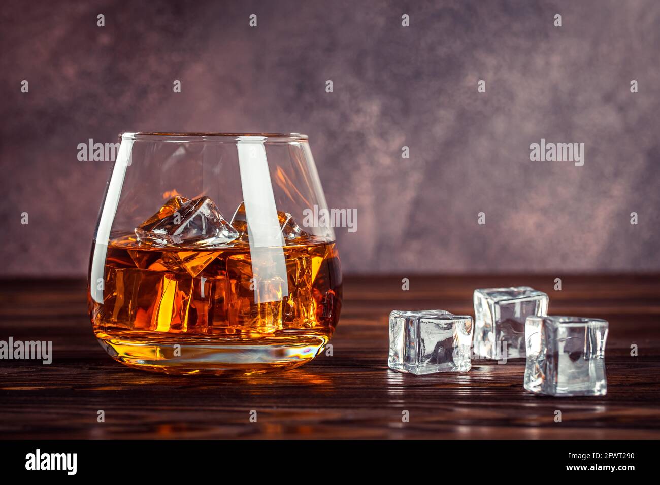Ein Glas Whiskey aus nächster Nähe. Schnaps mit Eis auf einem braunen Holztisch. Cognac, Bourbon. Starkes alkoholisches Getränk. Transparentes, gelbes alkoholisches Getränk Stockfoto