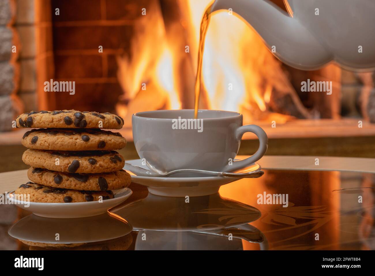 Gemütliche Kamin-Szene, Tee fließt in weiße Tasse, Kekse auf einem Teller, Reflexion von einem Feuer auf einem Glastisch. Stockfoto