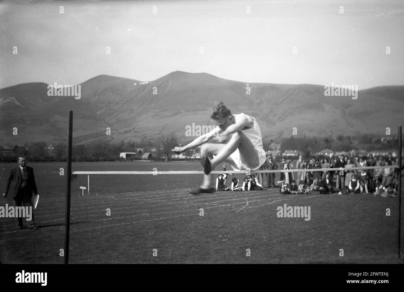 1951, historisches Bild, vor dem Hintergrund von Bergketten, draußen auf einem Grasfeld, ein Schuljunge beim Hochsprung, Highlands, Schottland. Beobachtet von einer Menge Eltern und Kindern, ist seine Technik beim Versuch, über die horizontale Stange zu springen, ein traditioneller Sprung mit beiden Füßen nach vorne. Stockfoto