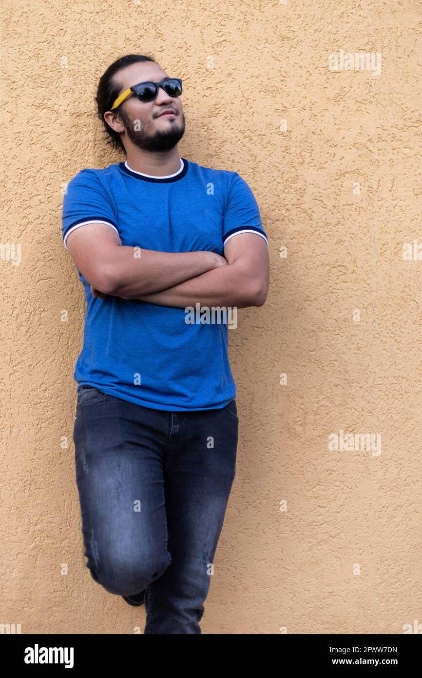 Junger Latino-Mann mit Sonnenbrille und gekreuzten Armen steht in einer senffarbenen Wand und blickt zum Horizont. Vertikale Aufnahme. Frisches Konzept Stockfoto