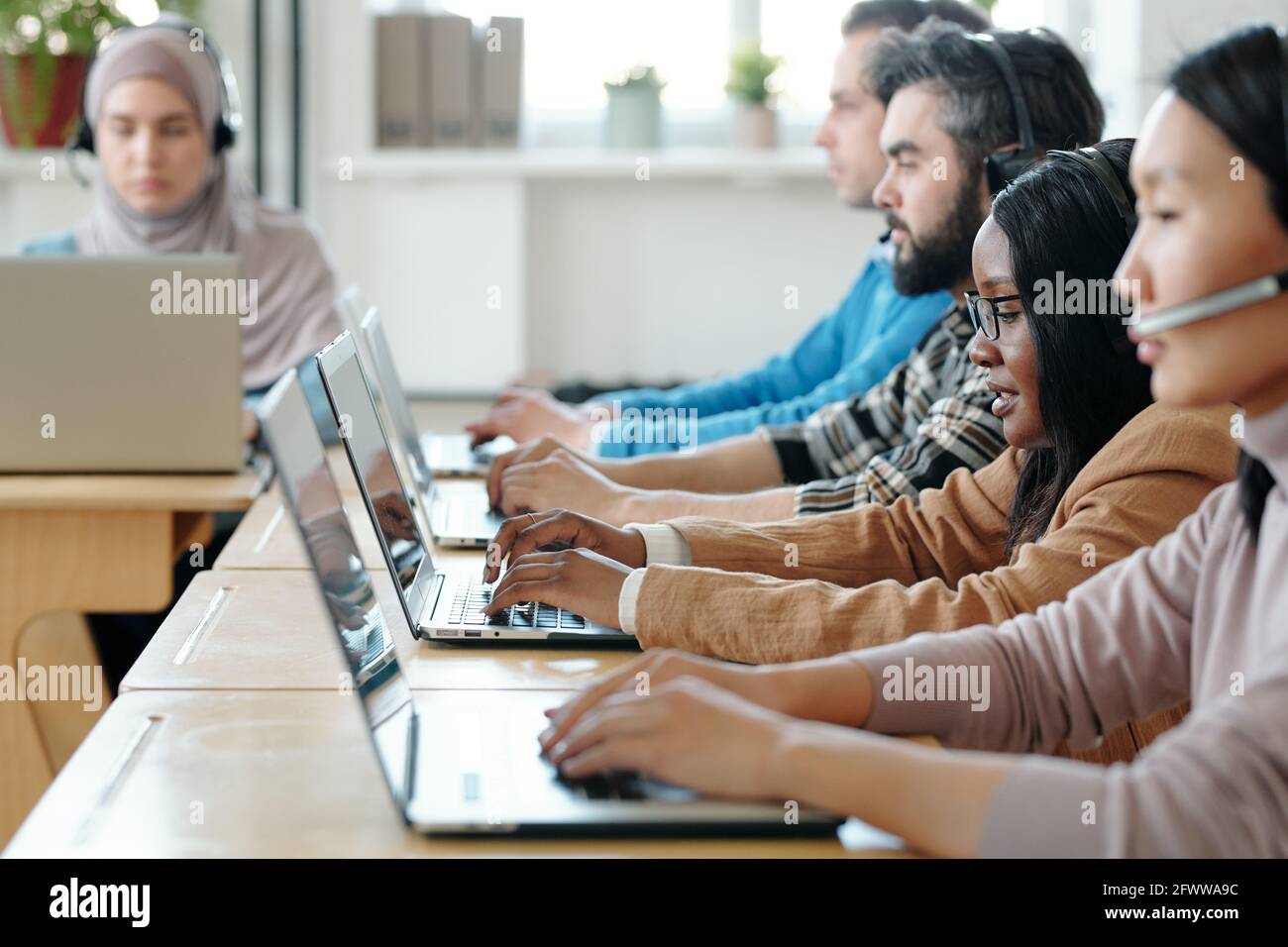 Reihe von seriös multiethnischen Support-Personal an Schreibtischen sitzen und Tippen Sie auf Laptops, während Sie Anrufe über schnurlose Headsets mit entgegennehmen Mikrofone Stockfoto