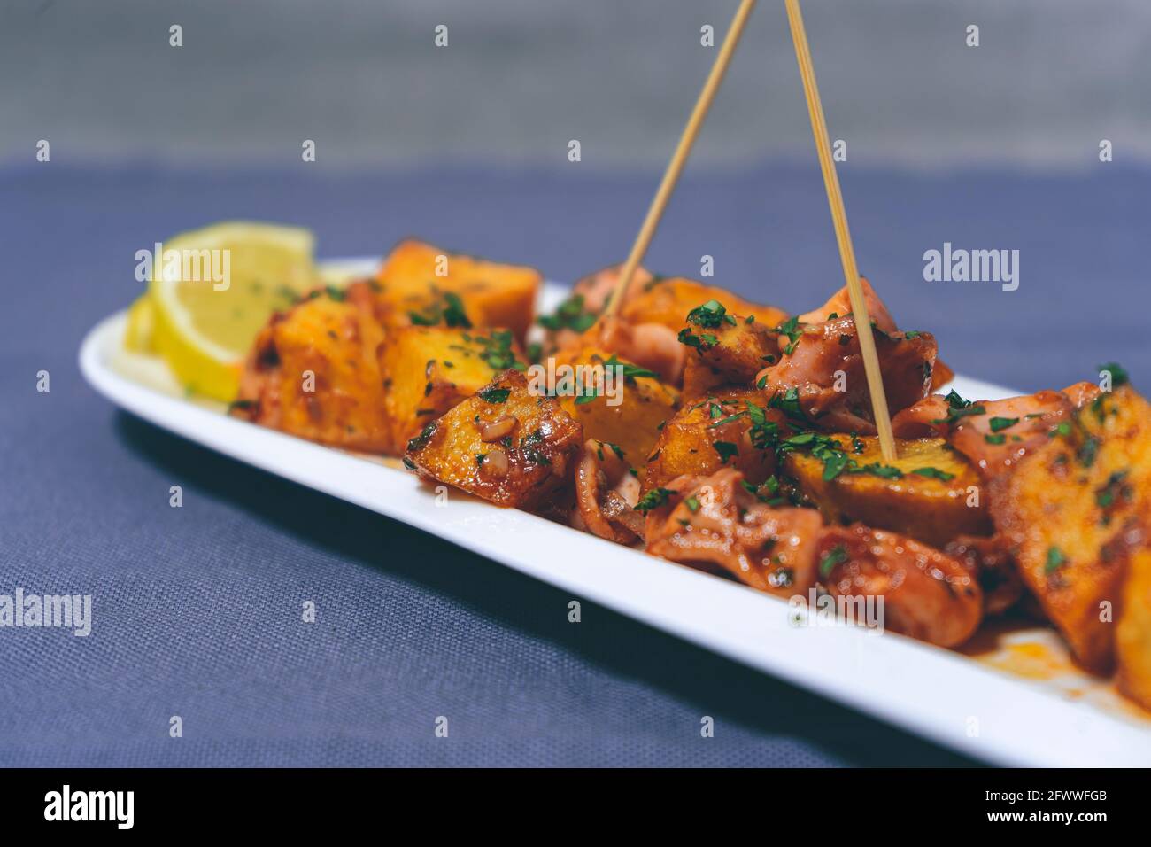 Aperitif oder typisch spanische Tapa, Tintenfisch mit Knoblauch und Kartoffeln, serviert auf einem länglichen weißen Tablett mit Zitrone und rustikalem Hintergrund. Traditionell Stockfoto