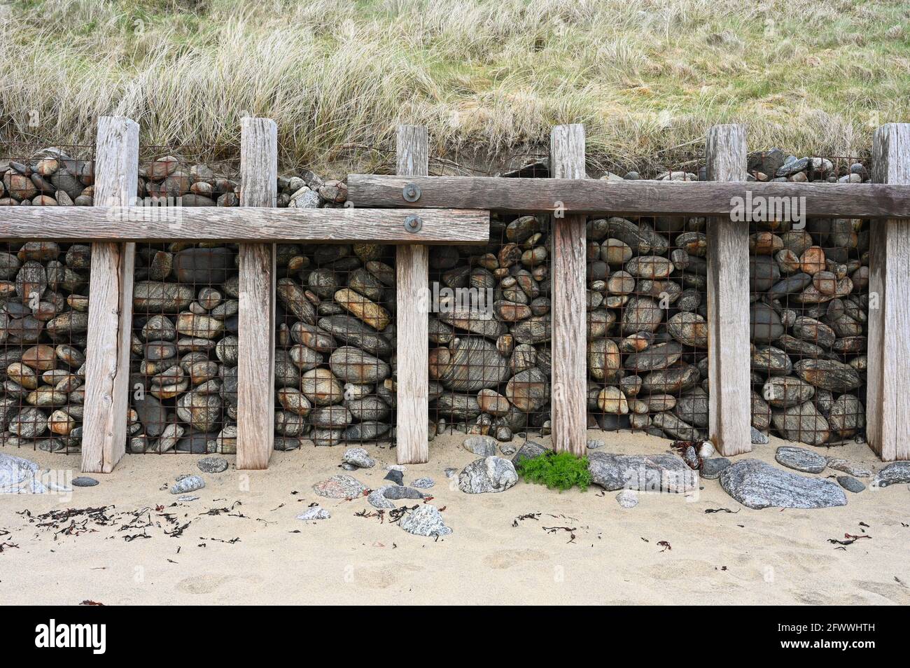 Küstenmauer aus Stein, Metallgeflecht und Holzsäulen. Sand im Vordergrund, Dünengras über der Wand. Stockfoto