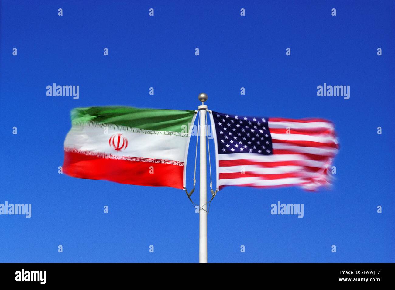 Iranische und amerikanische Flaggen schwenken Seite an Seite in gegenläufigen Winden auf einem einzigen Fahnenmast. Digitales Composite. Stockfoto