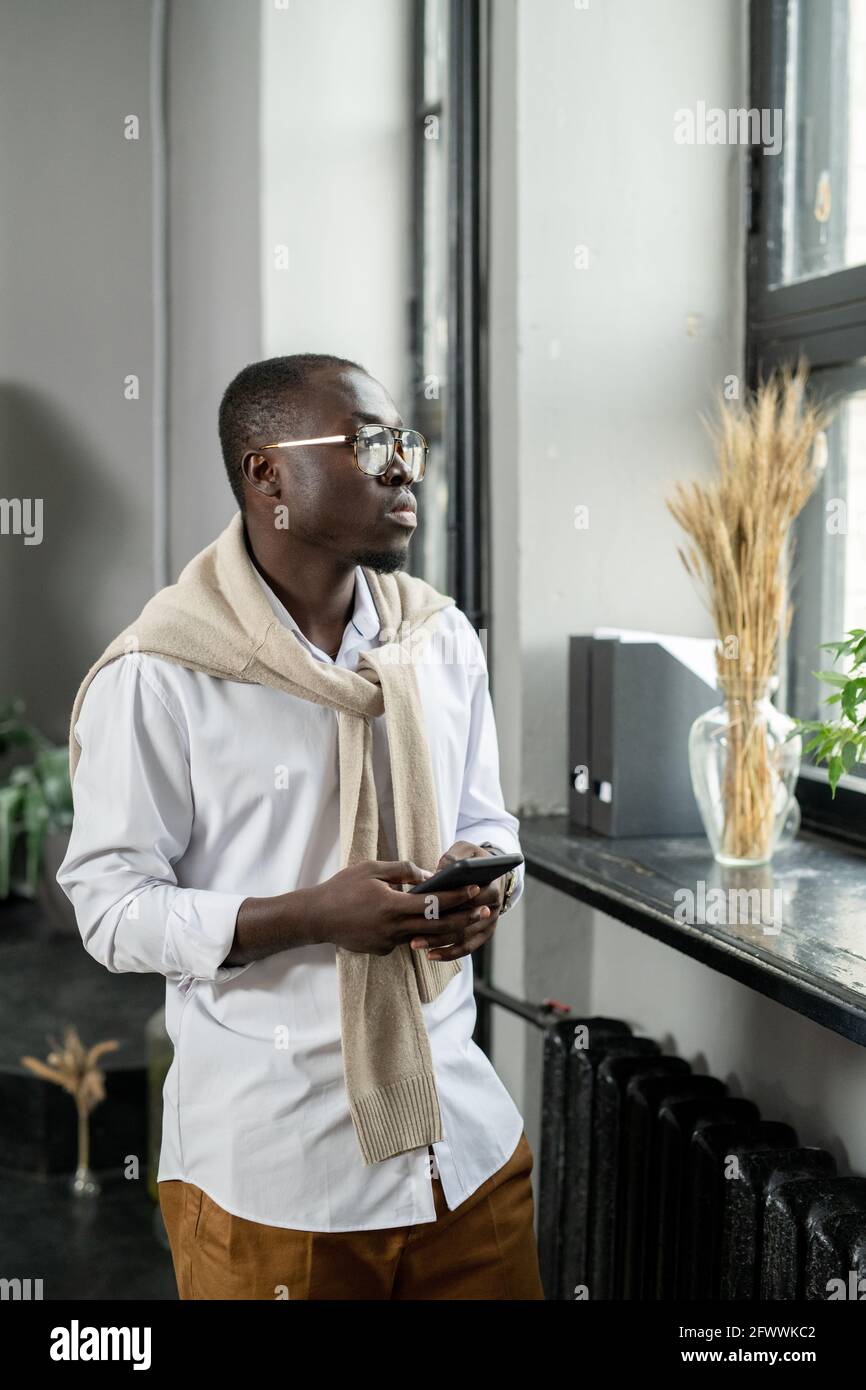 Junger afrikanischer Geschäftsmann in formalwear scrollt im Smartphone und sucht Durch das Fenster Stockfoto