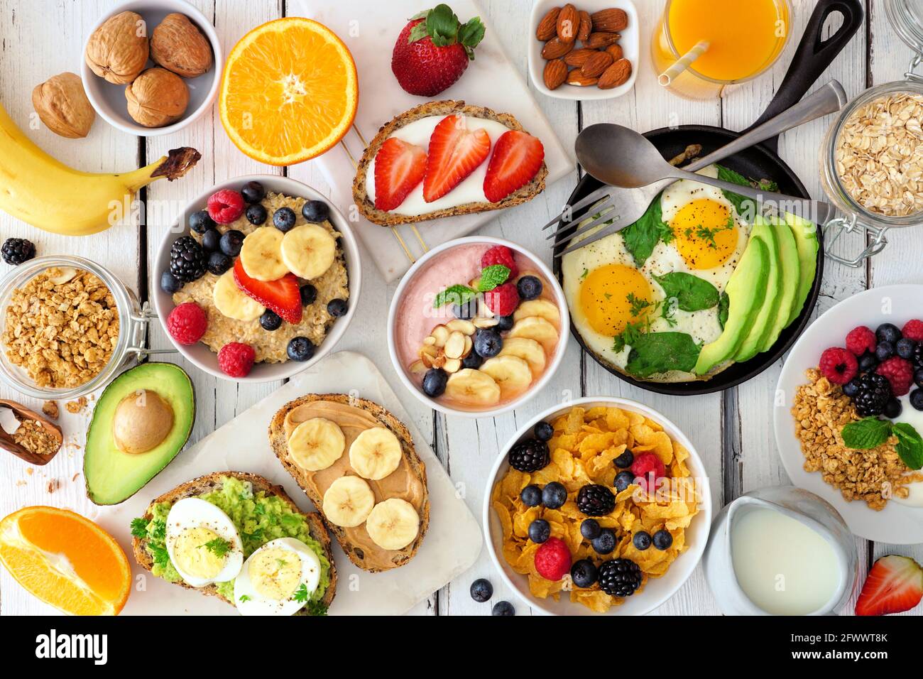 Gesunde Frühstückstisch-Szene mit Obst, Joghurt, Haferflocken, Smoothie-Schüssel, Müsli, Nahrhafte Toast und Eierpfanne. Blick von oben auf einen weißen Holzhinterteil Stockfoto