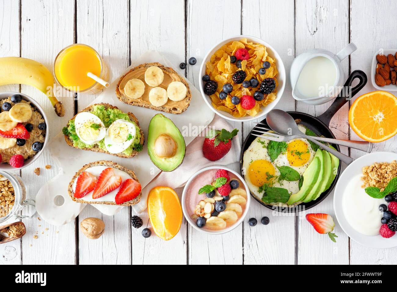 Gesunde Frühstückstisch Szene mit Obst, Joghurt, Haferflocken, Müsli, Smoothie Schüssel, Nahrhafte Toast und Eierpfanne. Blick von oben auf eine weiße Holzrückseite Stockfoto