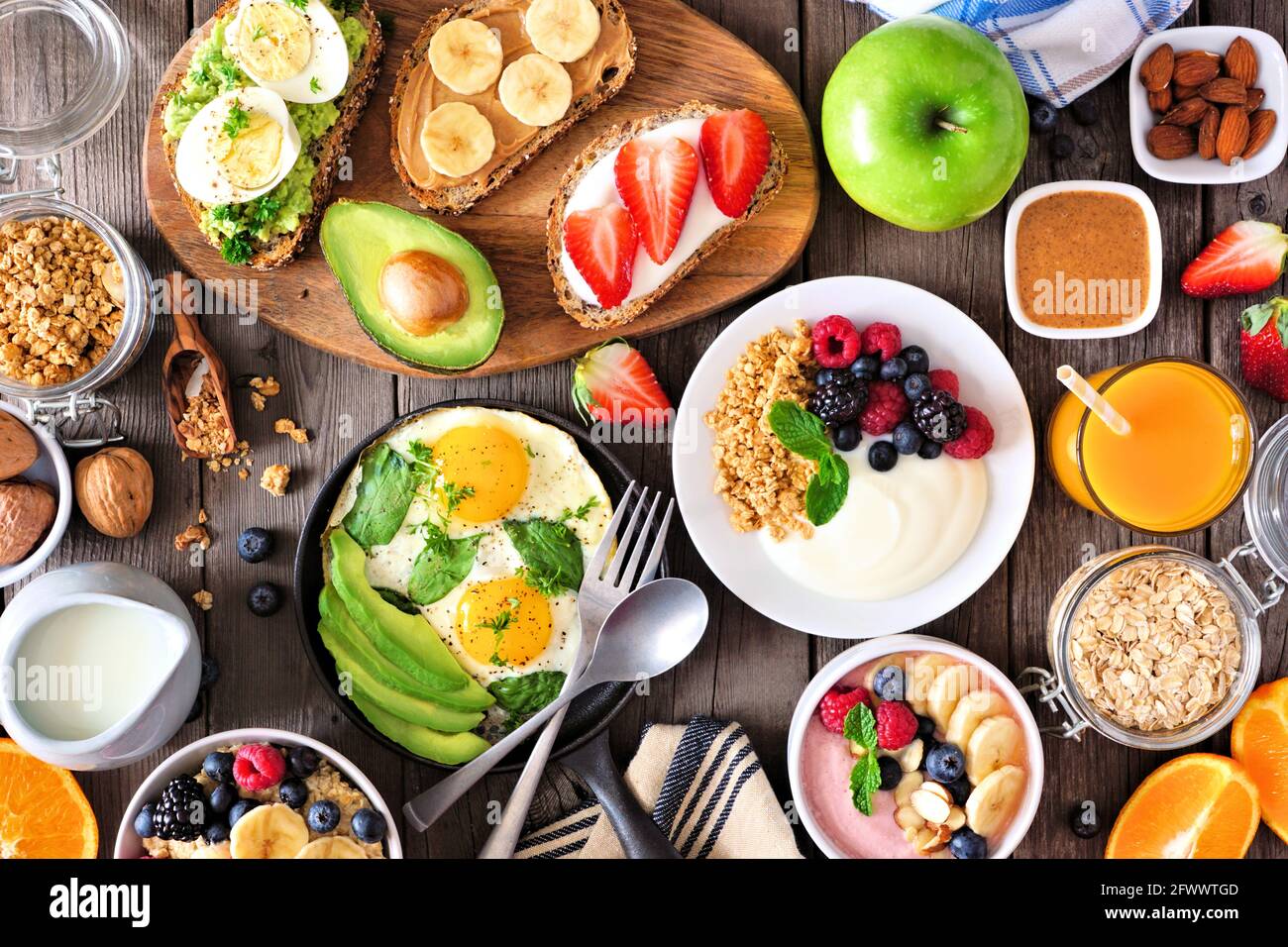 Gesunde Frühstückstisch-Szene mit Obst, Joghurt, Smoothie-Schüssel, Haferflocken, nahrhaftem Toast und Eierpfanne. Blick über einen Holzhintergrund. Stockfoto