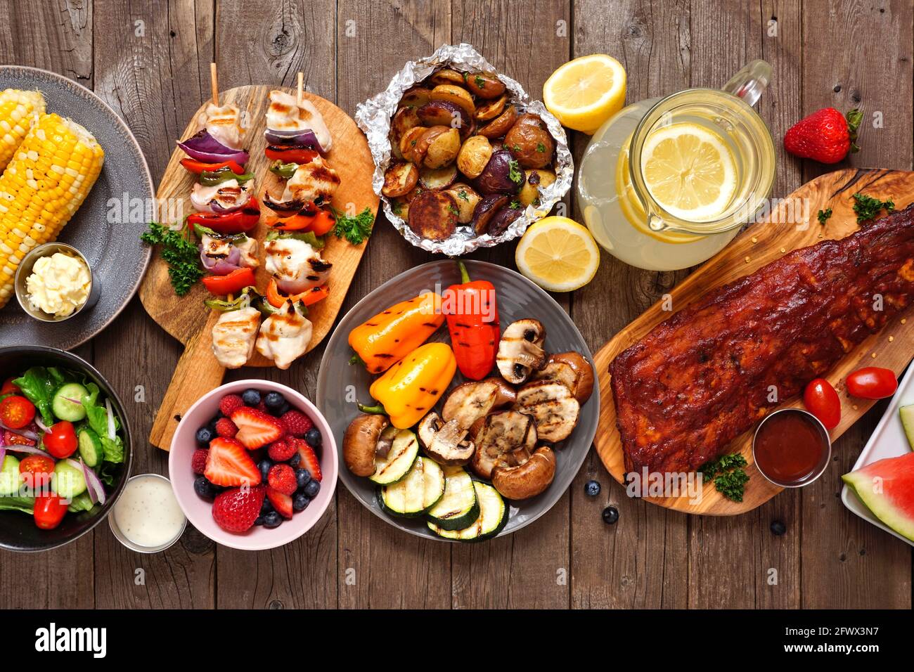 Sommer-BBQ oder Picknick-Tischszene. Auswahl an gegrilltem Fleisch, Gemüse, Obst, Salat und Kartoffeln. Draufsicht über einen dunklen Holzhintergrund. Stockfoto
