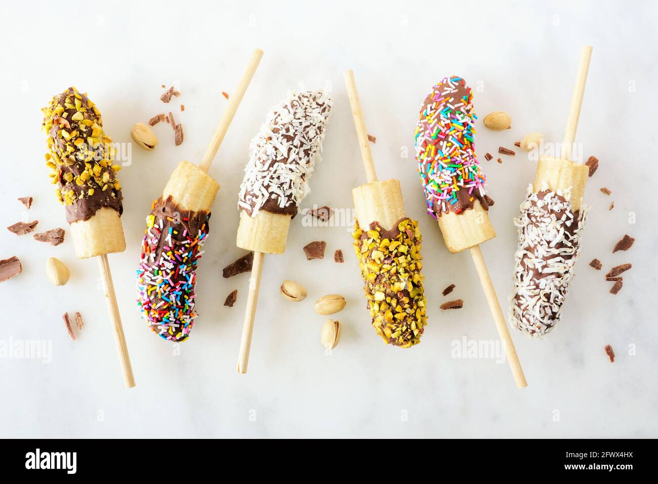 Gefrorene, in Schokolade getauchte Bananenpops mit Nüssen, Streuseln und Kokosnuss. Blick von oben auf weißem Marmor Hintergrund verstreut. Stockfoto