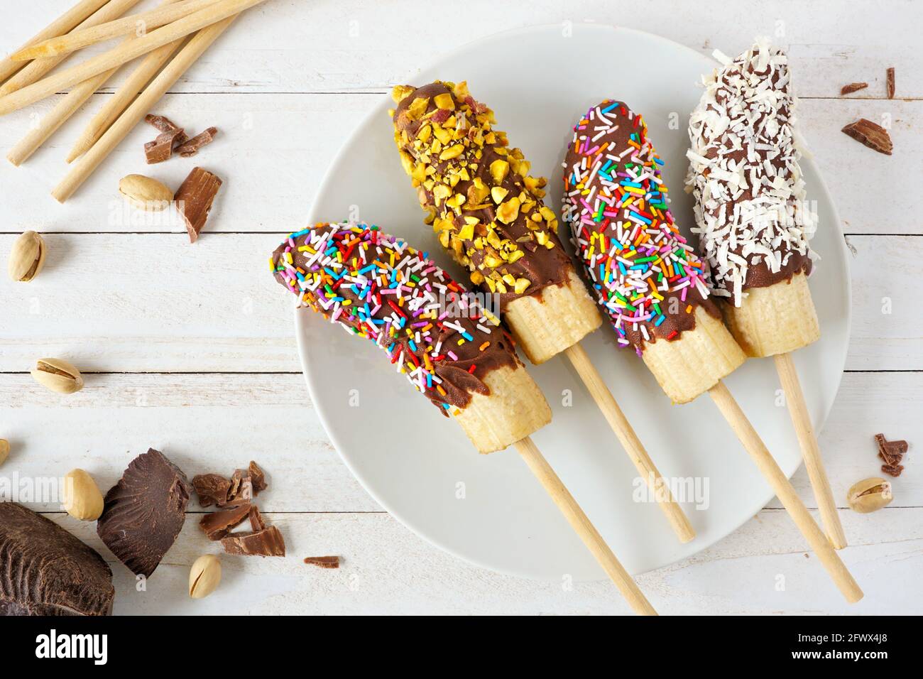 Gefrorene, in Schokolade getauchte Bananenpops mit Streuseln, Nüssen und Kokosnuss. Deckenansicht Tischszene auf einem Holzhintergrund. Stockfoto