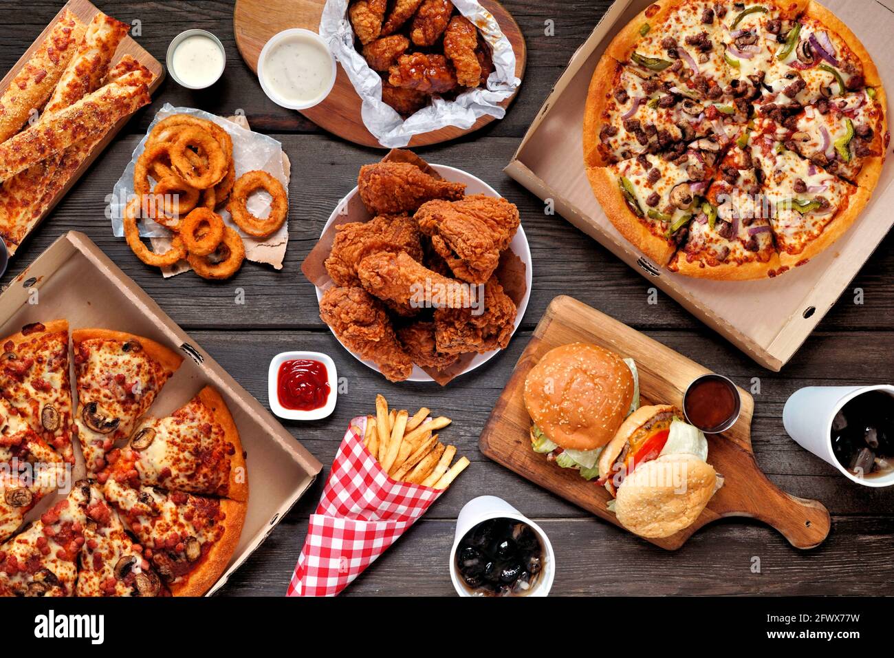 Buffet-Tisch mit Speisen zum Mitnehmen oder Anliefern. Pizza, Hamburger, gebratenes Huhn und Beilagen. Oben auf einem dunklen Holzhintergrund. Stockfoto