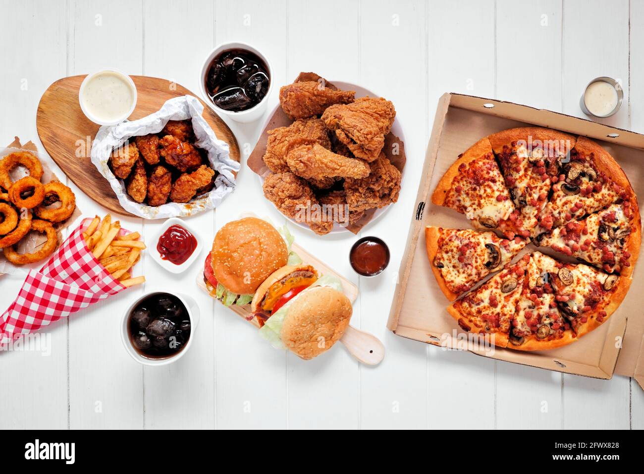 Tischszene mit großer Auswahl an Take-Out- und Fast-Food-Gerichten. Hamburger, Pizza, gebratenes Huhn und Beilagen. Draufsicht auf weißem Holzhintergrund. Stockfoto