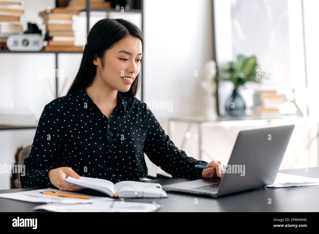 Erfolgreiche, schöne, junge asiatische Frau, Freiberuflerin oder Studentin, die am Schreibtisch sitzt, einen Laptop für die Arbeit oder das Online-Studium nutzt, sich während des Webinars oder Online-Vortrags Notizen macht und freundlich lächelt Stockfoto