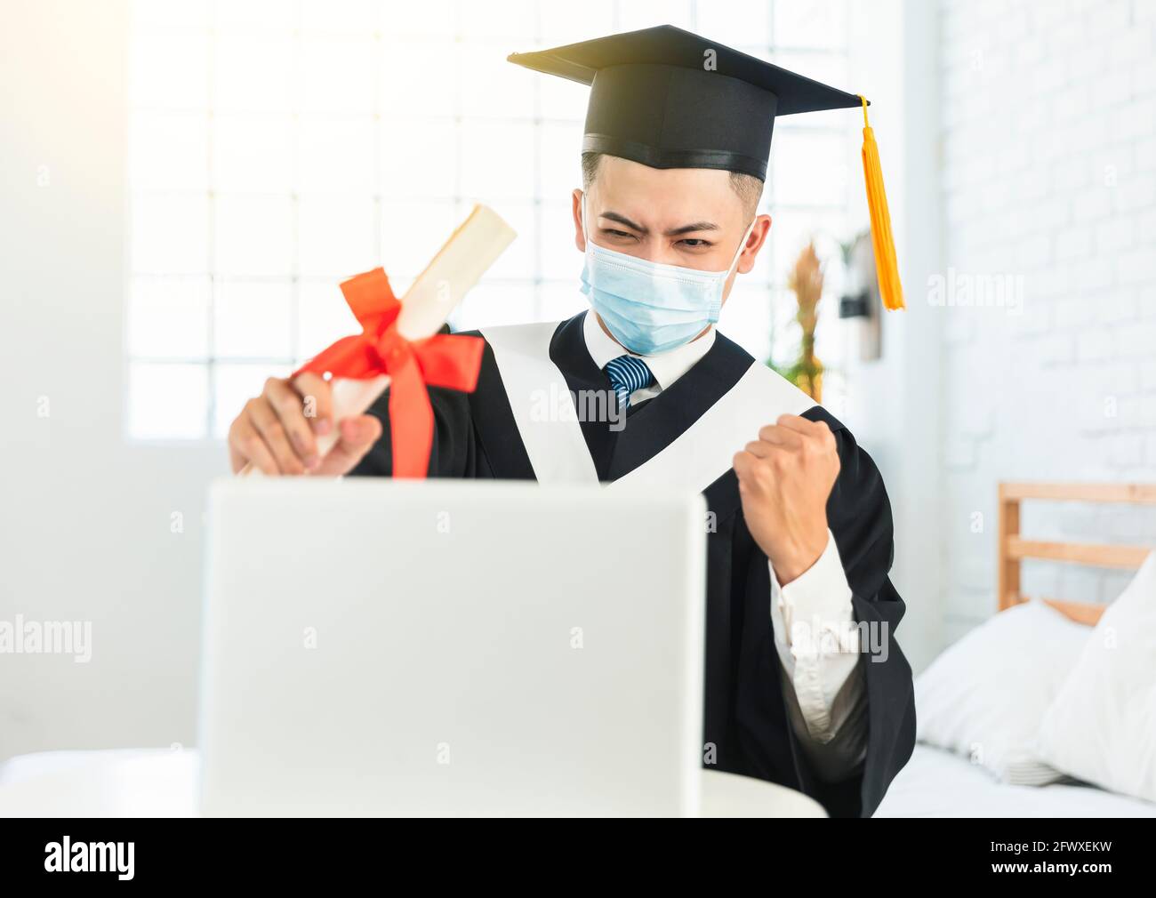 Glücklicher Student in medizinischer Maske und sitzt am Schreibtisch mit Laptop, hält Diplom und feiert Erfolg Stockfoto