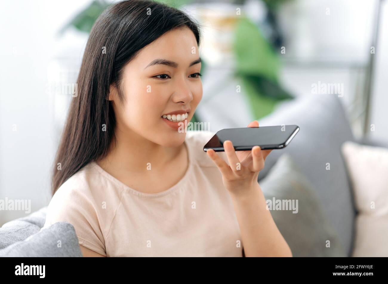 Junge glückliche schöne positive asiatische langhaarige Mädchen, sitzen auf dem Sofa, tragen T-Shirt, mit Smartphone, Chat mit Freunden oder Familie auf Freisprecheinrichtung oder Aufnahme von Audio-Nachricht, lächelt freundlich Stockfoto