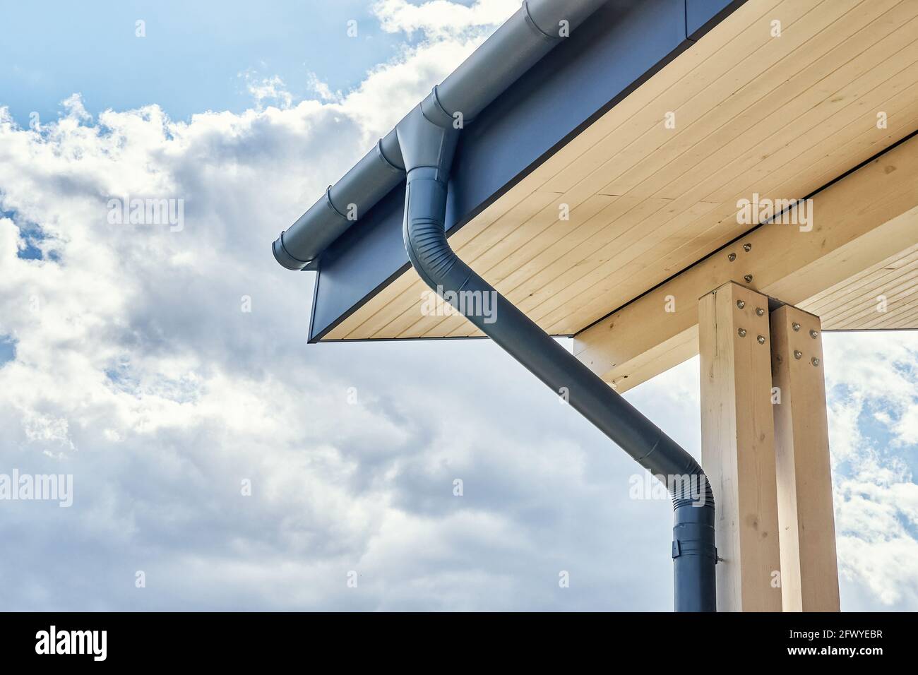 Moderne Regenwasserleitung aus grauem Metall auf dem Dach von New installiert Gebäude mit Holzterrasse am bewölkten Tag aus der Nähe Stockfoto