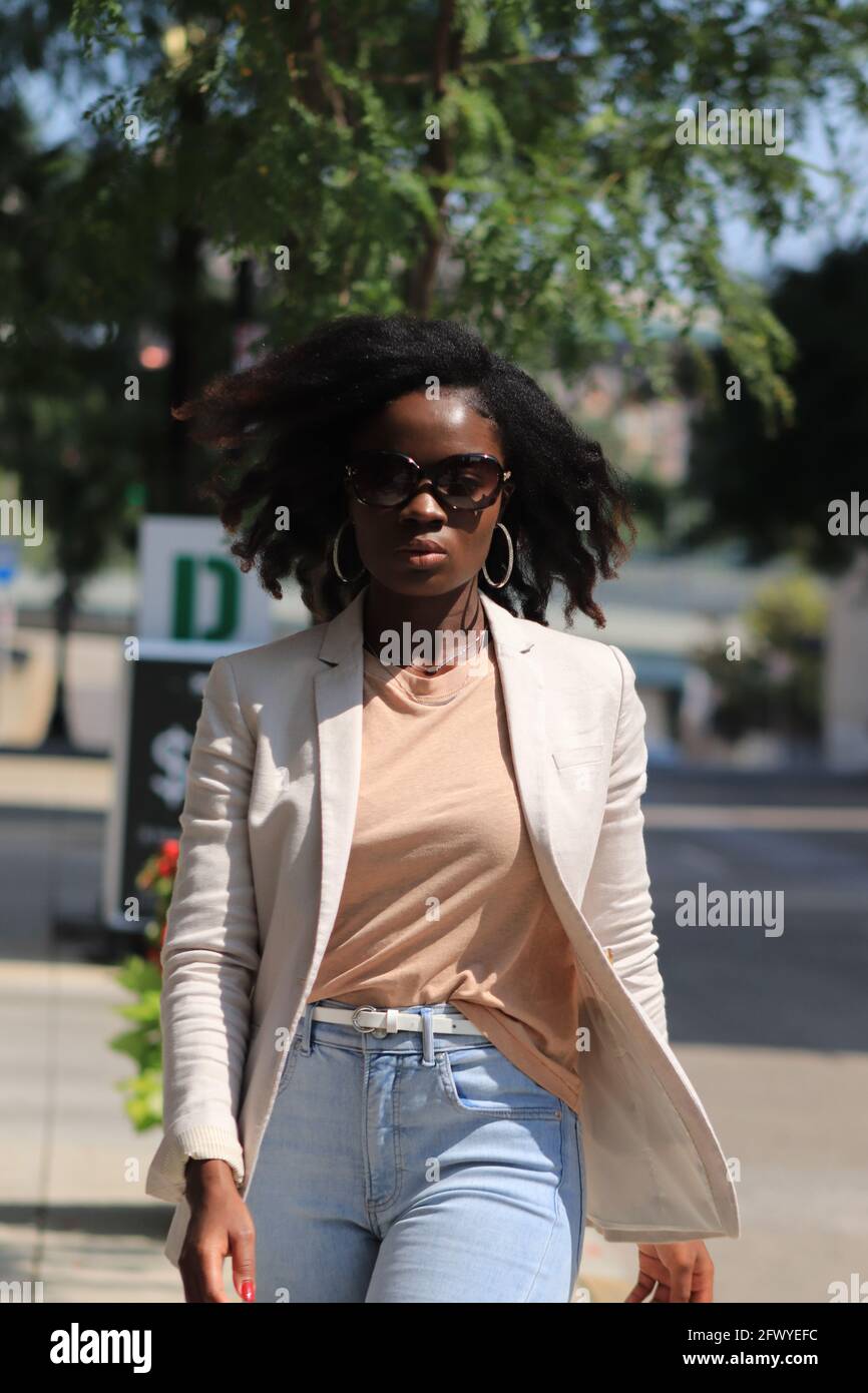 Schillerndes Model, das mit einer Sonnenbrille die Straße entlang geht. Nahaufnahme Porträt, afroamerikanisches Model, natürliche Frisur, Handtasche auf der Schulter, lässiger Blazer Stockfoto