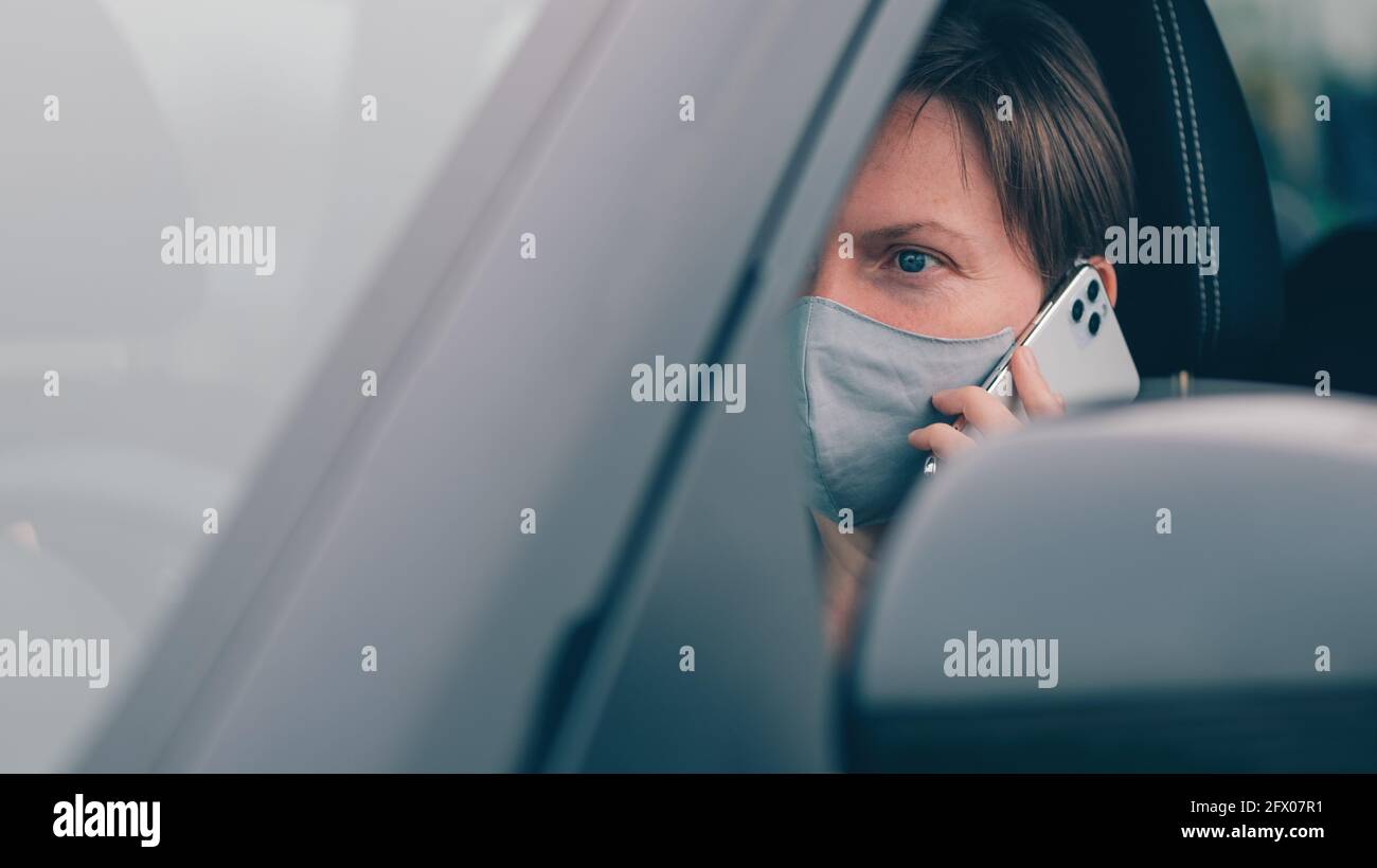 Erwachsene Frau mit schützender Gesichtsmaske, die während der Fahrt mit Covid-19-Pandemien von ihrem Auto aus auf dem Mobiltelefon spricht, selektiver Fokus Stockfoto