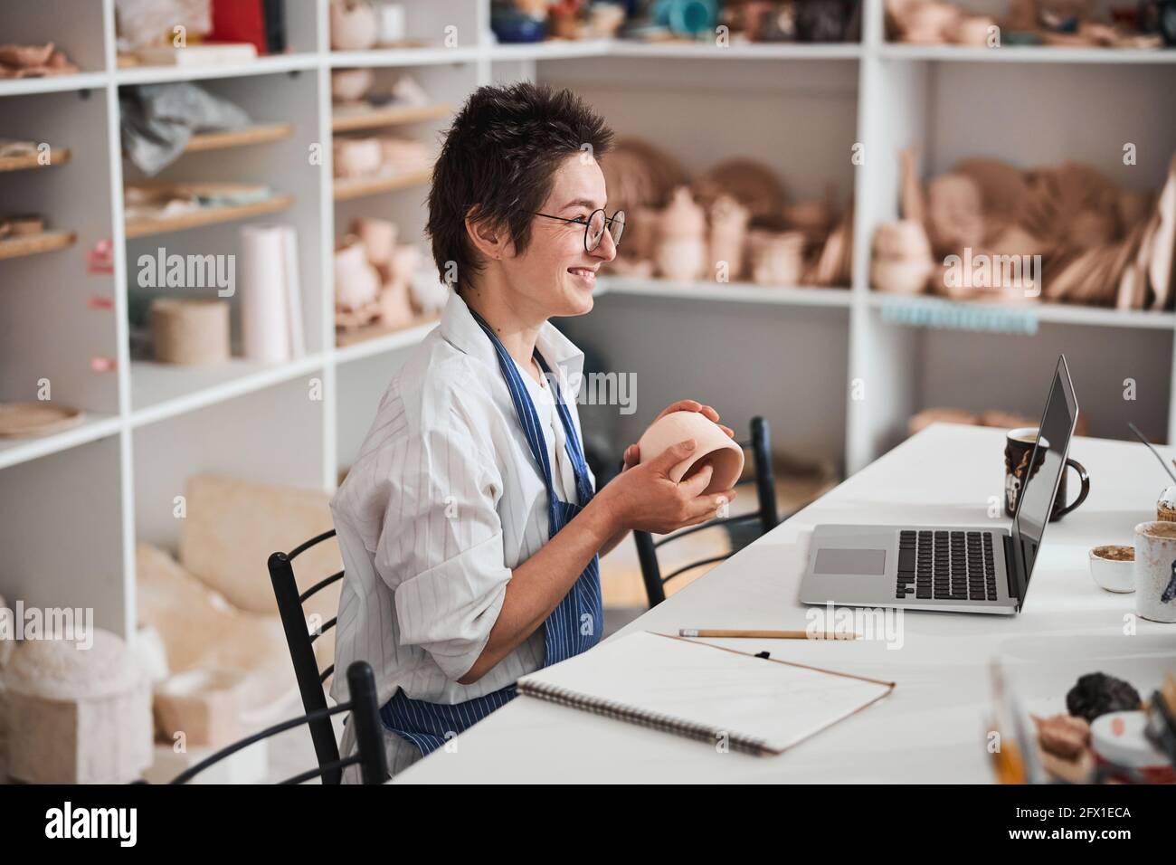 Überglücklich weibliche Keramiker kippte Tonbecher vor Laptop-Bildschirm Stockfoto