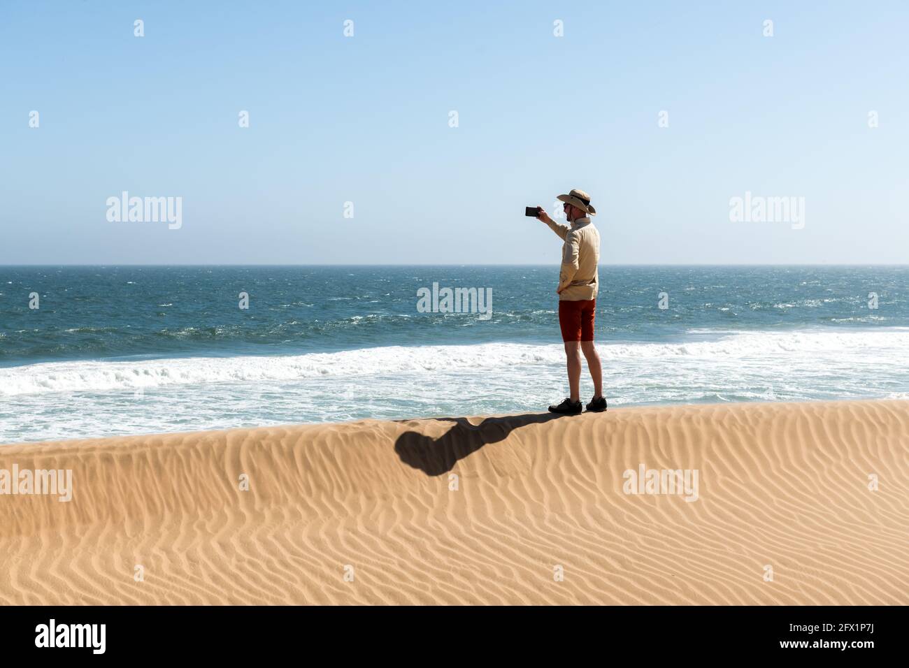 Ein einzelner Mann in einem Cowboyhut nimmt ein Selfie in der Namib-Wüste auf dem Hintergrund des Atlantiks auf. Reisekonzept Stockfoto