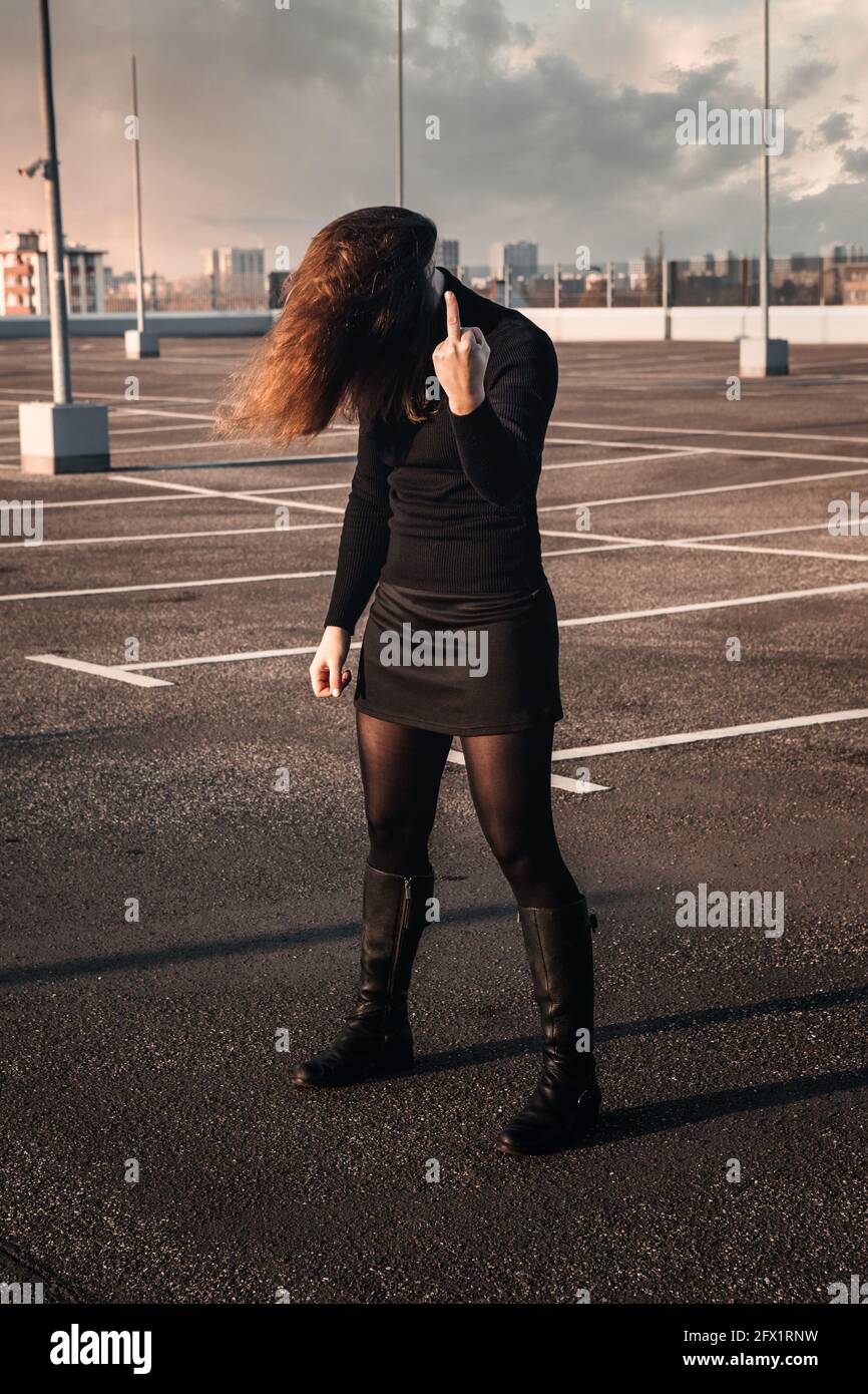 Brunette Frau steht auf leerem Parkplatz mit ihren Haaren Das Gesicht abdecken und den Mittelfinger zeigen Stockfoto