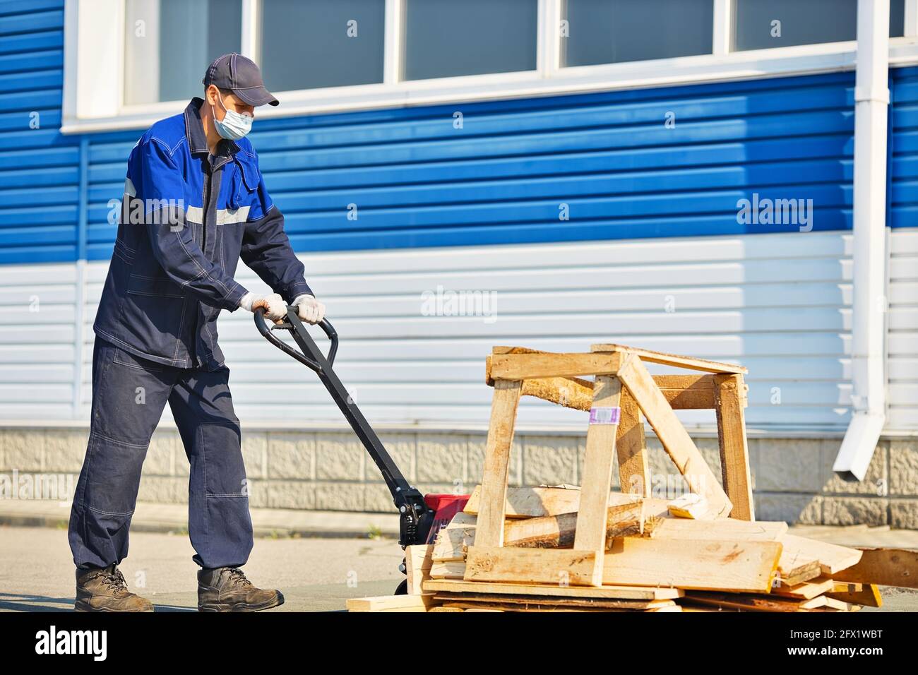 Ein Mann in Arbeitskleidung und einer medizinischen Maske trägt an einem Sommertag Holzbretter auf einem Wagen auf dem Territorium eines Industriestützpunktes. Arbeiten Sie gemäß den hygienischen Standards Stockfoto