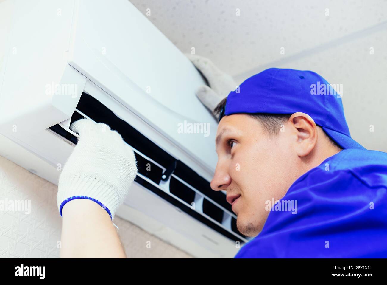 Ein junger Arbeiter mit blauer Baseballmütze und Handschuhen inspiziert und wartet die Klimaanlage im Zimmer. Porträt eines Technikers bei der Arbeit. Nahaufnahme. Modell Stockfoto