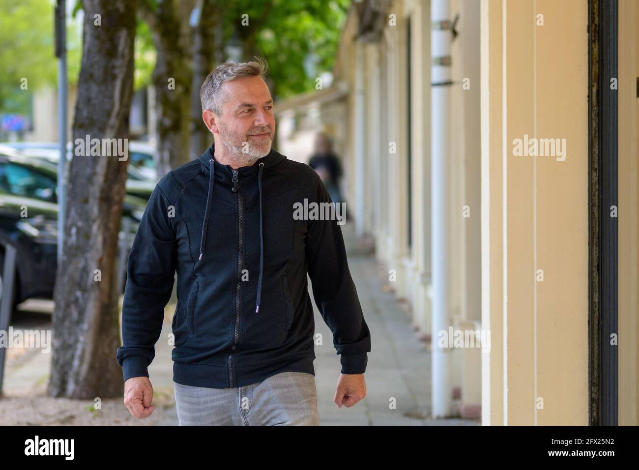 Ein Mann mittleren Alters in Freizeitkleidung, der eine urbane Straße entlang geht und sucht Mit einem Lächeln auf die Seite, während er sich dem nähert Kamera Stockfoto