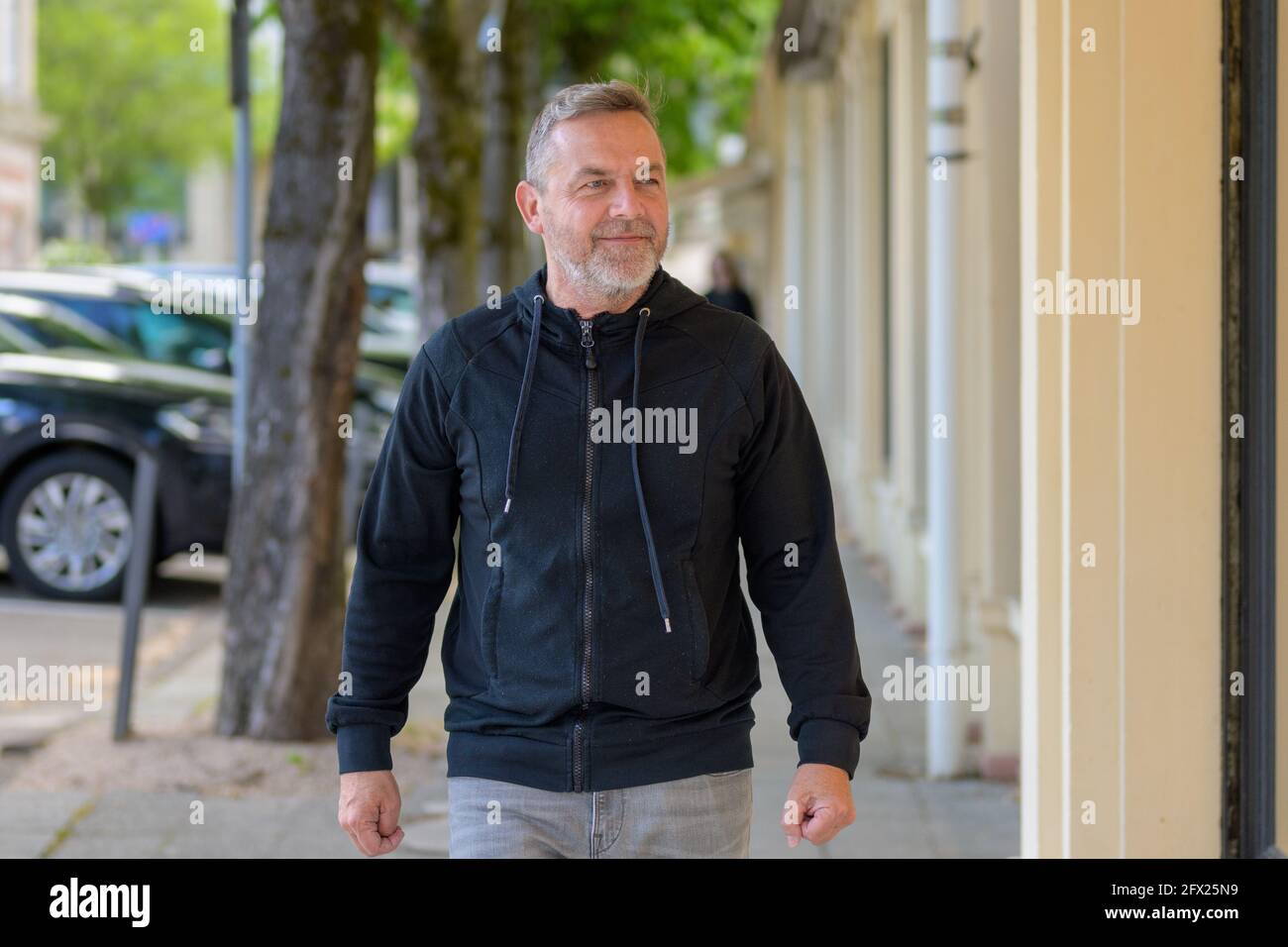 Ein Mann mittleren Alters in Freizeitkleidung, der eine urbane Straße entlang geht und sucht Mit einem Lächeln auf die Seite, während er sich dem nähert Kamera Stockfoto