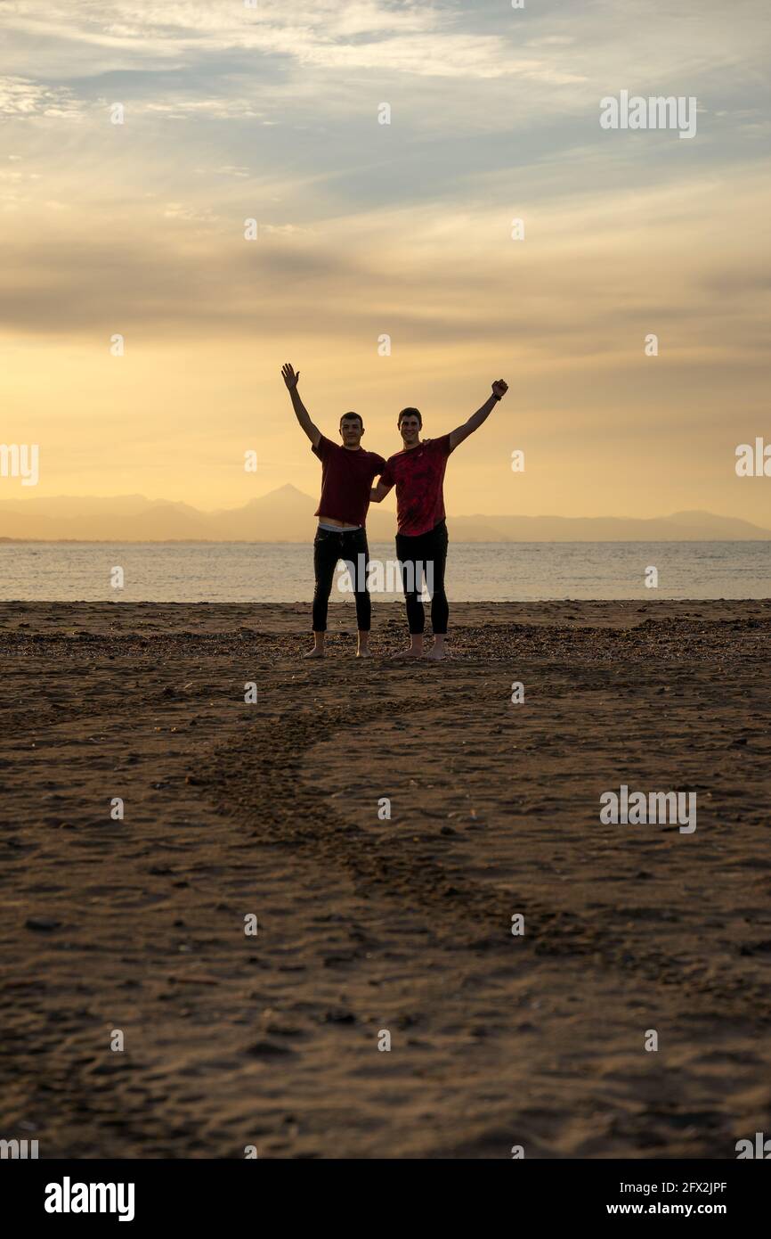 Zwei Silhouetten von Männern am Strand. In einem Sonnenuntergang mit einem ruhigen Meer.Freunde halten sich am Rücken und heben einen Arm vor Freude. Stockfoto