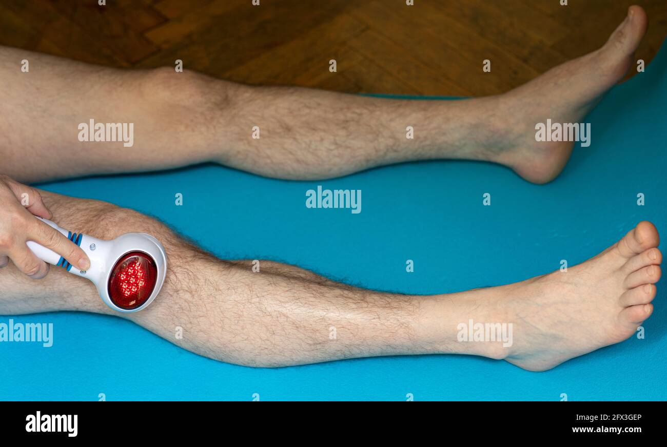 Der Mann massiert sein Knie mit einem elektrischen Massagegerät mit einem roten Licht der Hitze Stockfoto