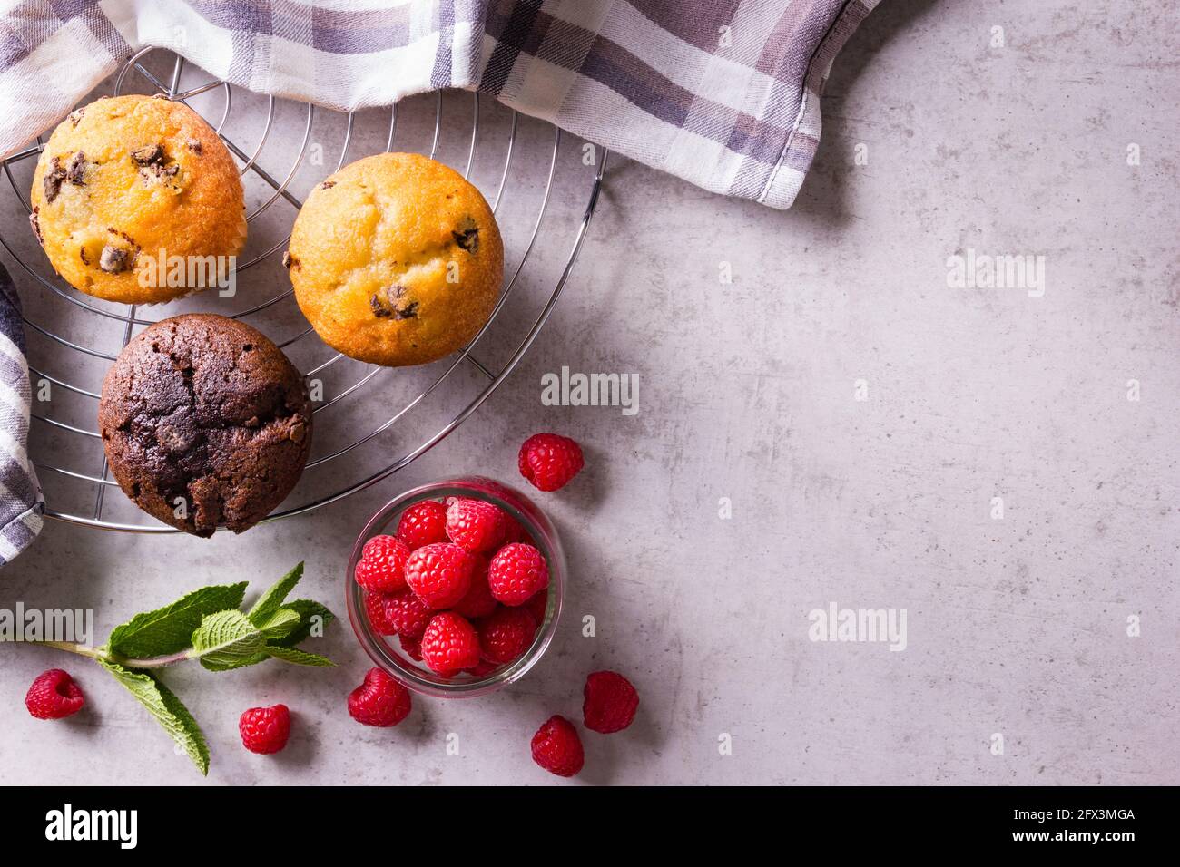 Frisch gebackene Cupcakes auf Kühlregal mit Serviette und Himbeeren Und frische Minze neben der grauen Arbeitsplatte Stockfoto