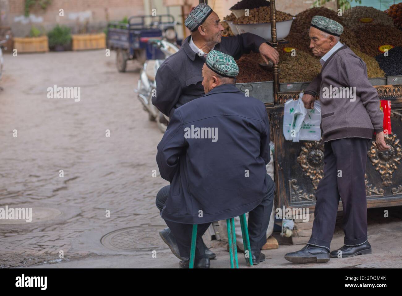 Drei alte Männer unterhalten sich neben einem Wagen getrockneter Früchte in Kahgar, Volksrepublik China Stockfoto