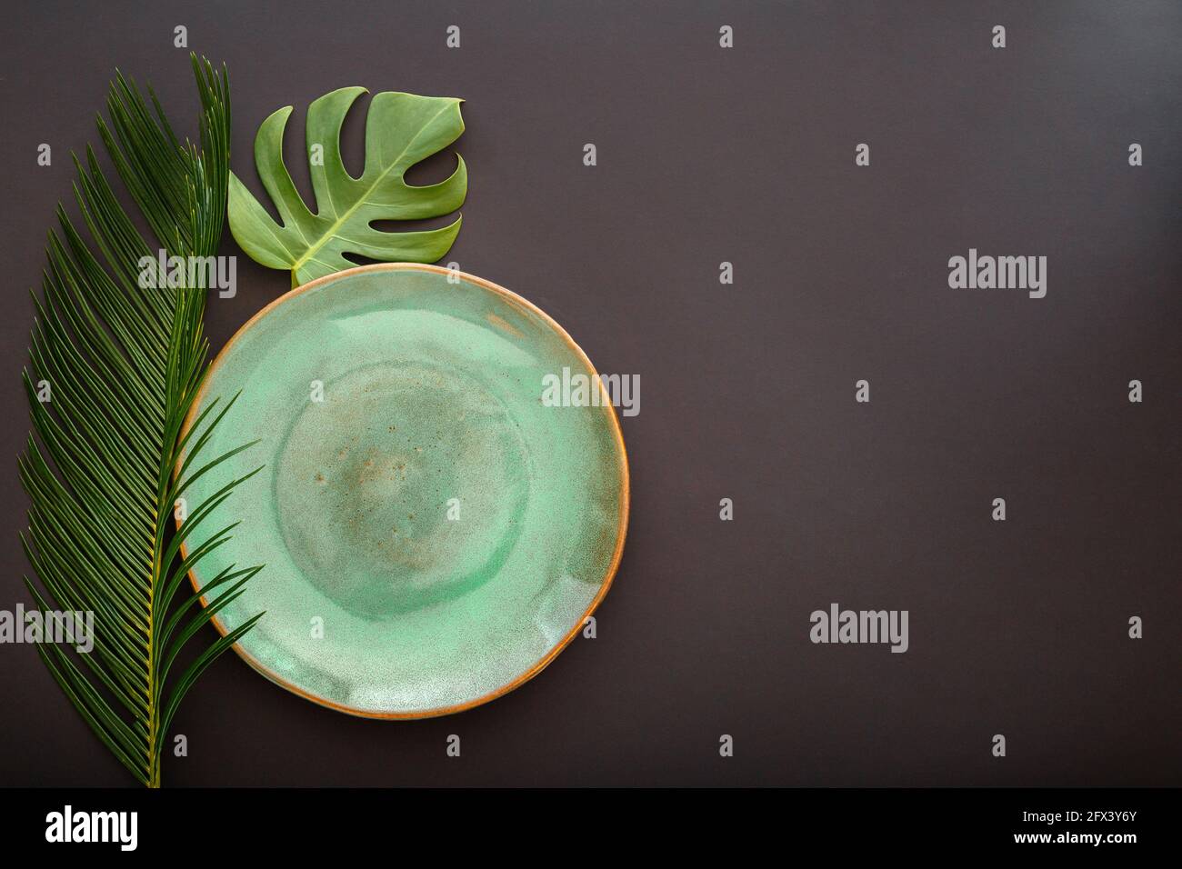 Leere grüne Platte auf schwarzem Hintergrund. Luxuriöser Speiseteller, serviert mit einer Monstera-Pflanze im asiatischen Kinfolk-Grunge-Stil mit tropischen Palmenblättern. Modell leer Stockfoto