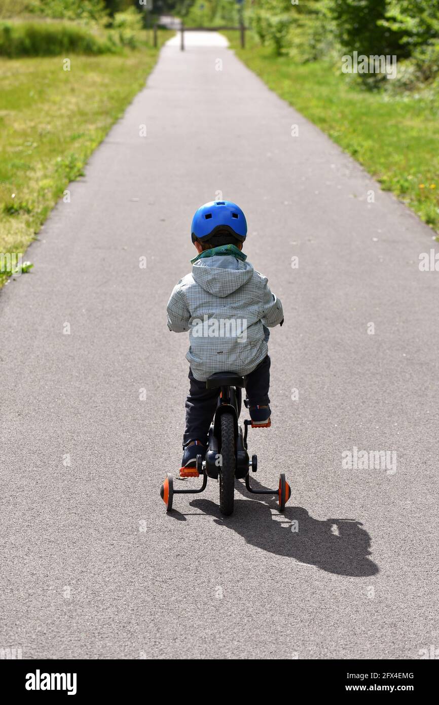 Rückansicht eines Kindes mit blauem Helm, das auf einer Straße ein Fahrrad mit Trainingsrädern fährt. Stockfoto