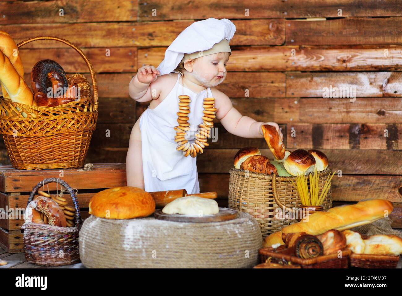 Ein kleiner Junge mit Kochmütze und Schürze und einem Haufen Bagels um den Hals sitzt vor dem Hintergrund von Backwaren in einem Brotladen. Stockfoto