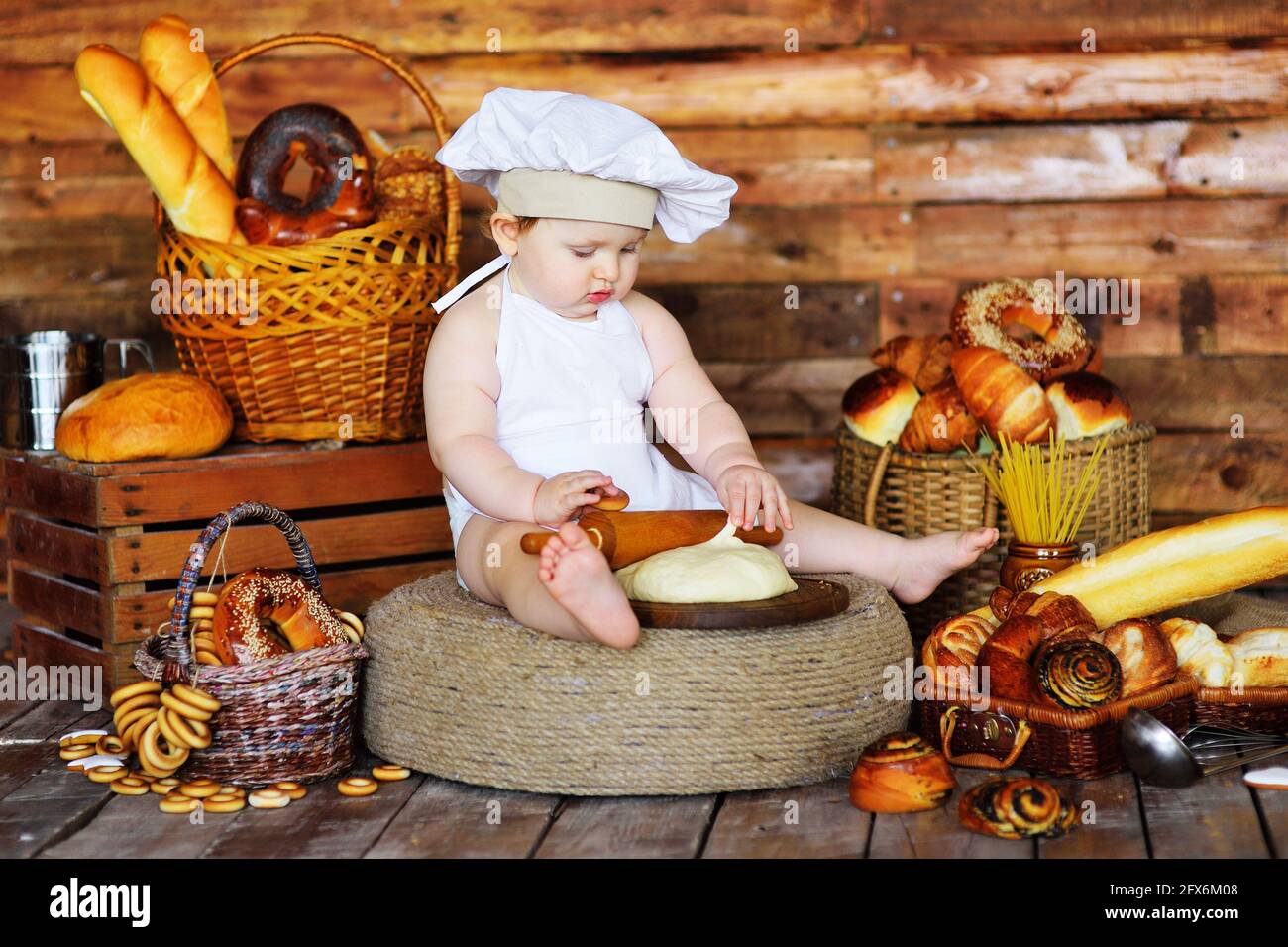 Der kleine Bäckerjunge mit Kochmütze und Schürze rollt rohen Teig mit einem hölzernen Nudelholz vor dem Hintergrund von Backwaren aus. Stockfoto