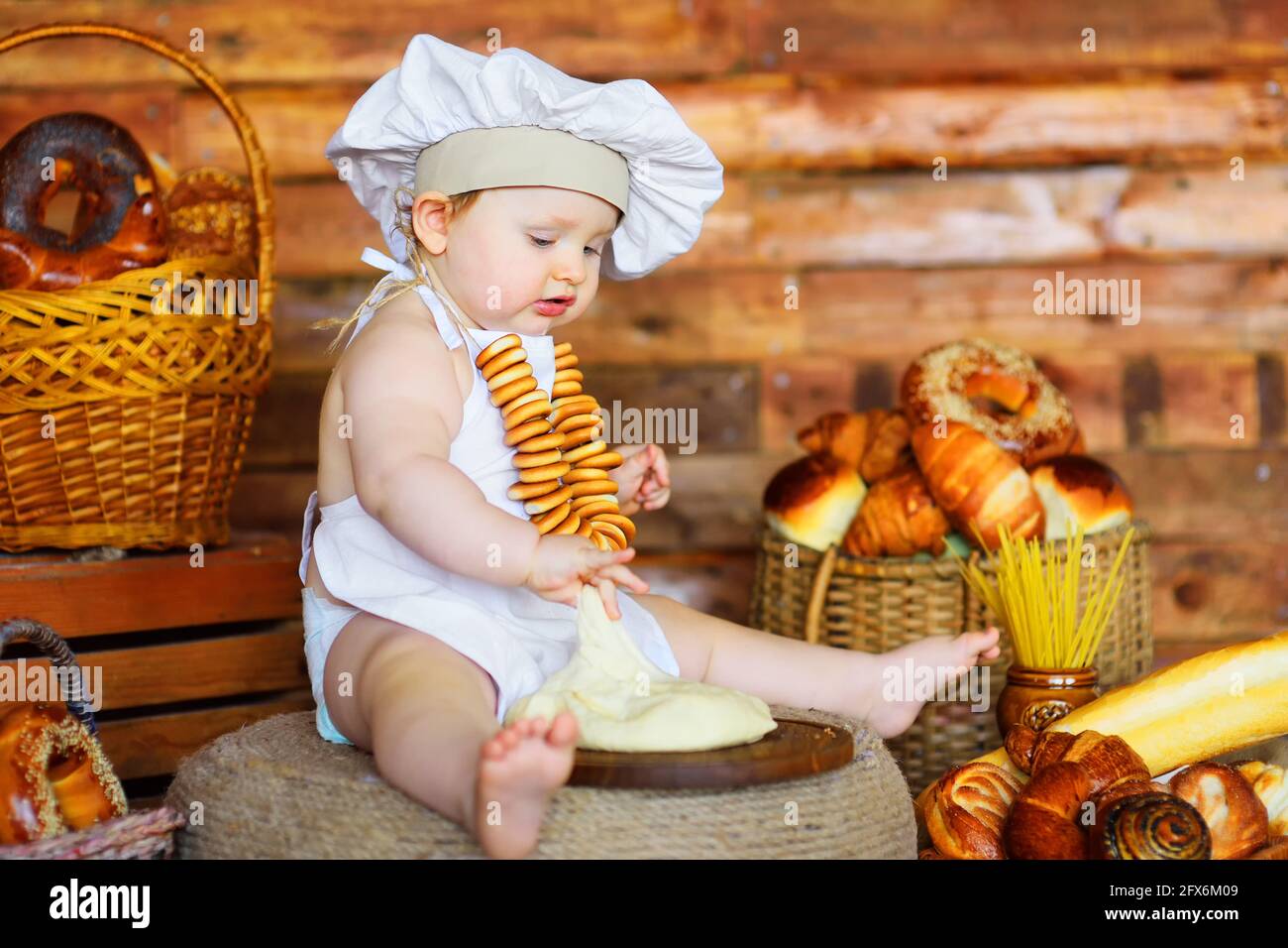 Der kleine Bäckerjunge mit Kochmütze und Schürze und einem Haufen Bagels um den Hals bereitet vor dem Hintergrund von Backwaren Teig zum Backen vor. Stockfoto