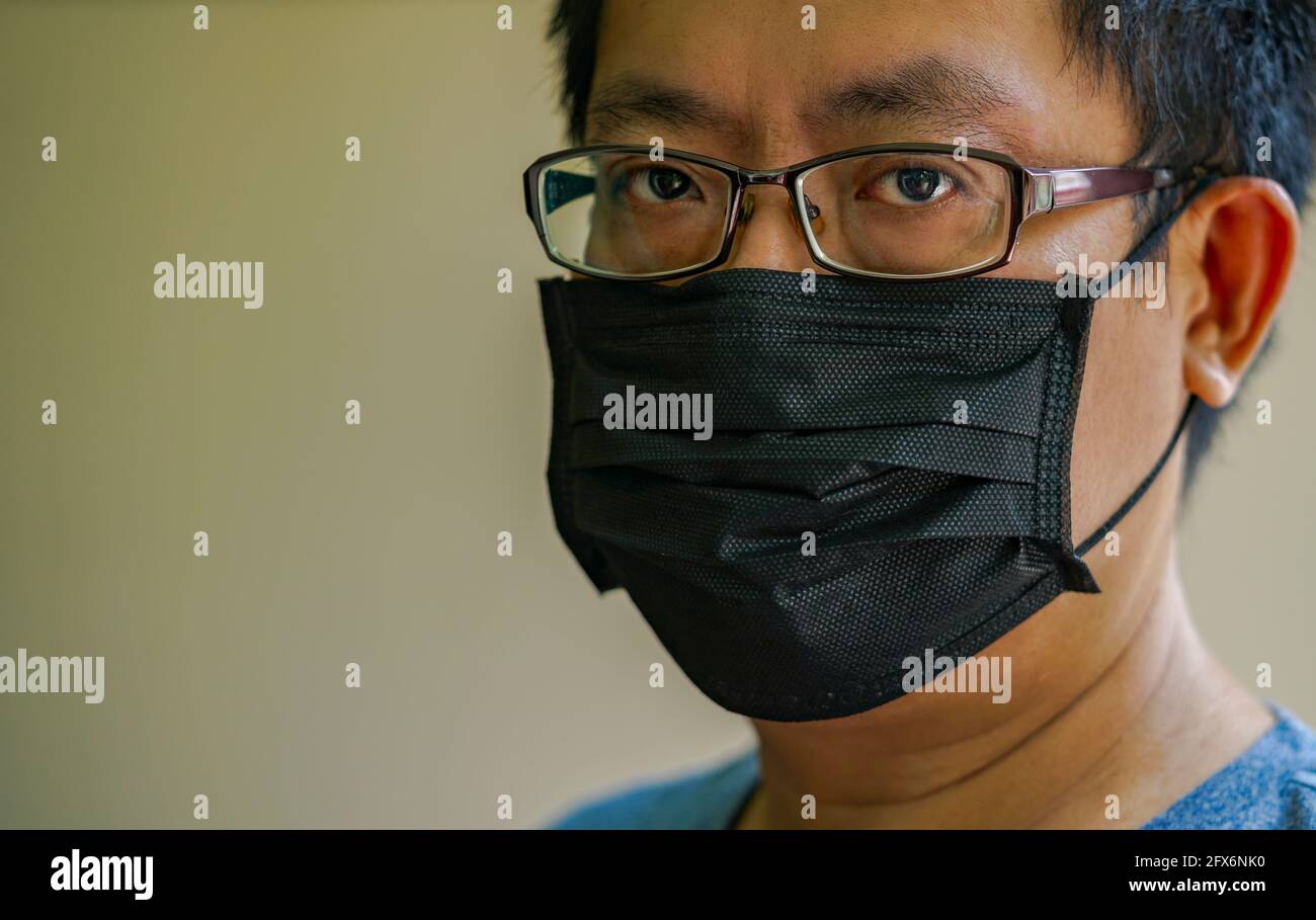 Nahaufnahme Mann Gesicht trägt Maske, um Virus-Epidemie zu schützen. Asiatischer Mann mittleren Alters mit Brille und schwarzer medizinischer Maske, Augen starren ernsthaft auf die Kamera. Stockfoto